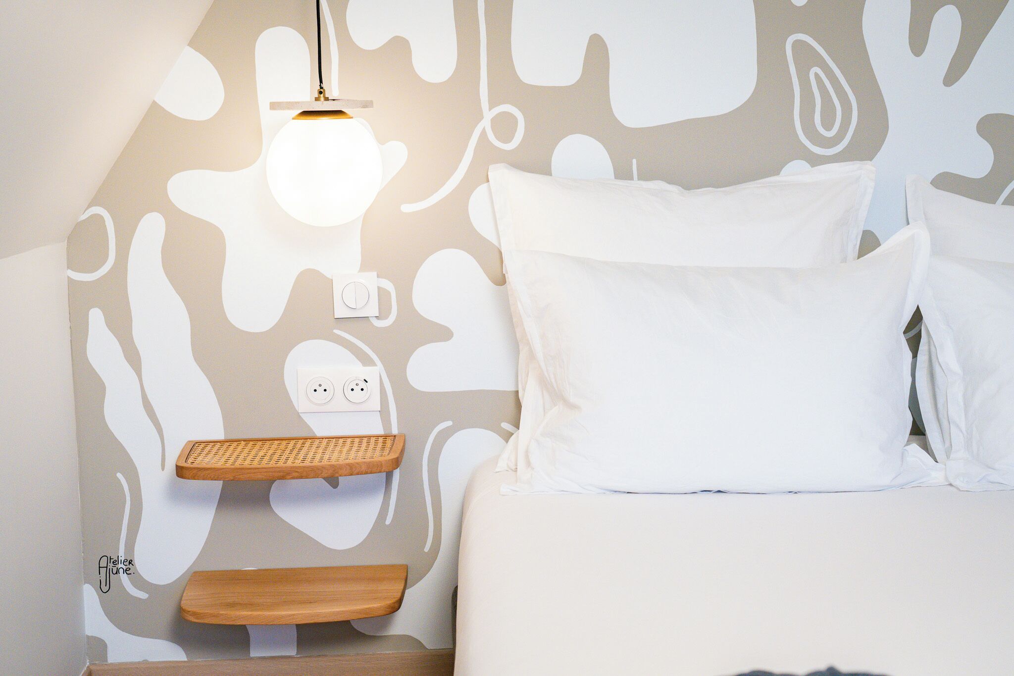 A bedroom with a bed, white pillows, a bedside table, a wall-mounted light fixture, and a power outlet.