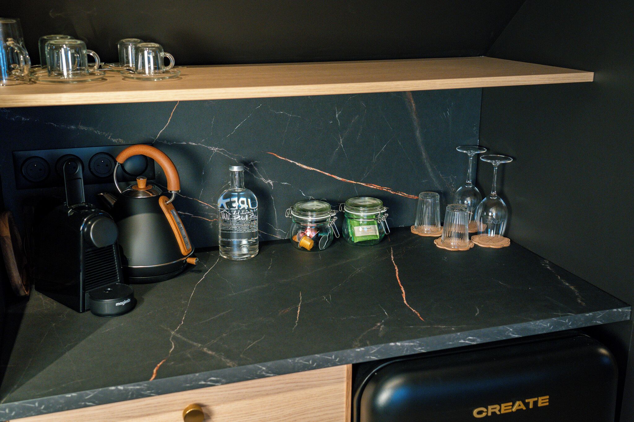 A kitchen counter with a coffee maker, a kettle, a bottle, jars, and wine glasses.