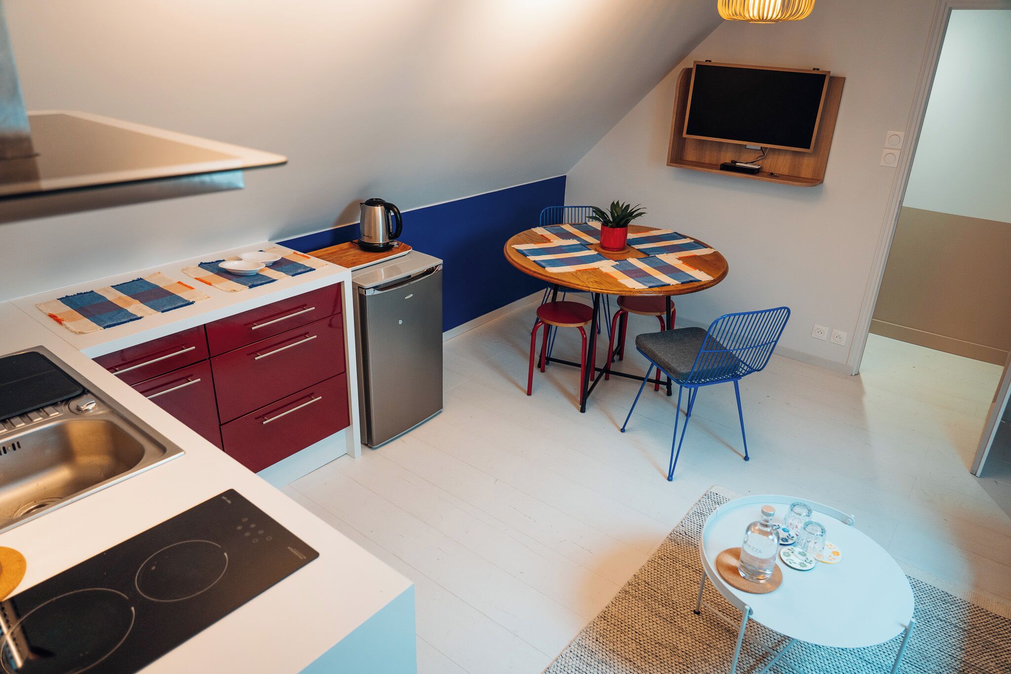 A compact kitchen with a stove, sink, and refrigerator, a small dining table with chairs, and a TV mounted on the wall.