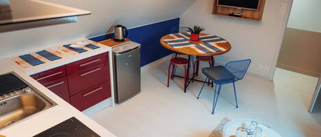 A compact kitchen with a stove, sink, and refrigerator, a small dining table with chairs, and a TV mounted on the wall.