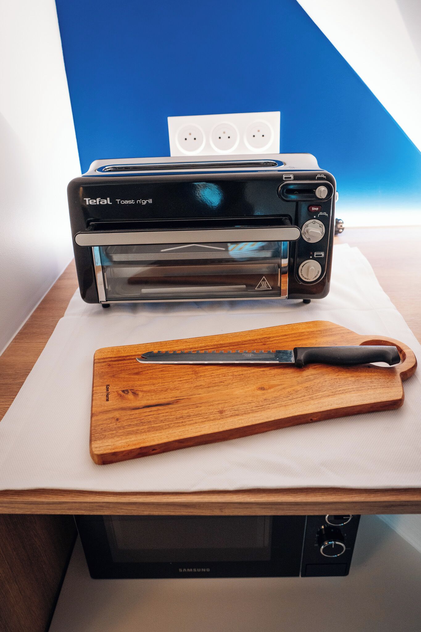 A toaster oven on a wooden cutting board with a knife, a Samsung microwave below, and a blue wall in the background.