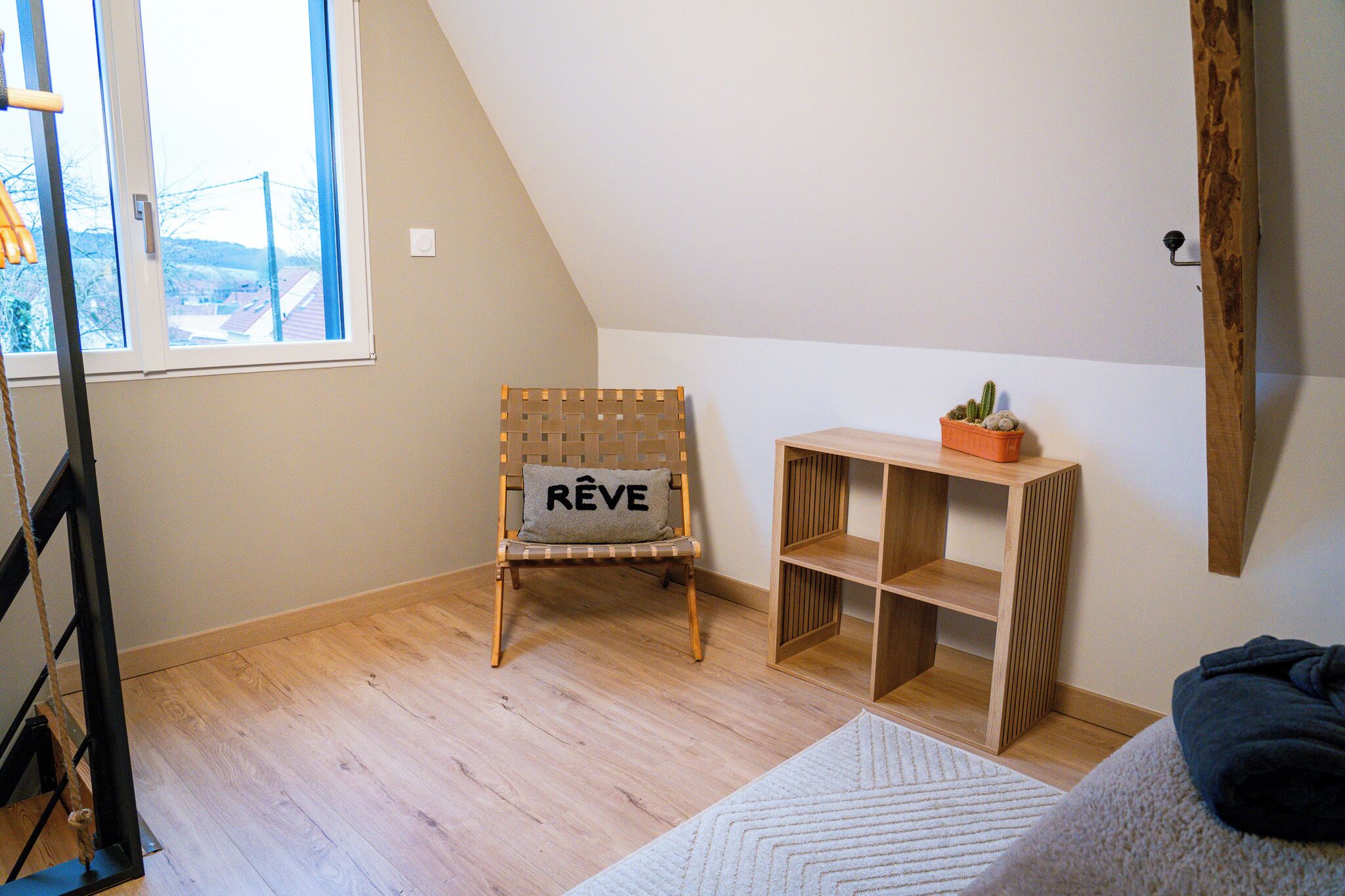Una habitación con techo inclinado, una silla de madera con un cojín que dice "REVE", un pequeño estante de madera con un cactus y una alfombra blanca.