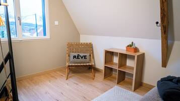 Una habitación con techo inclinado, una silla de madera con un cojín que dice "REVE", un pequeño estante de madera con un cactus y una alfombra blanca.