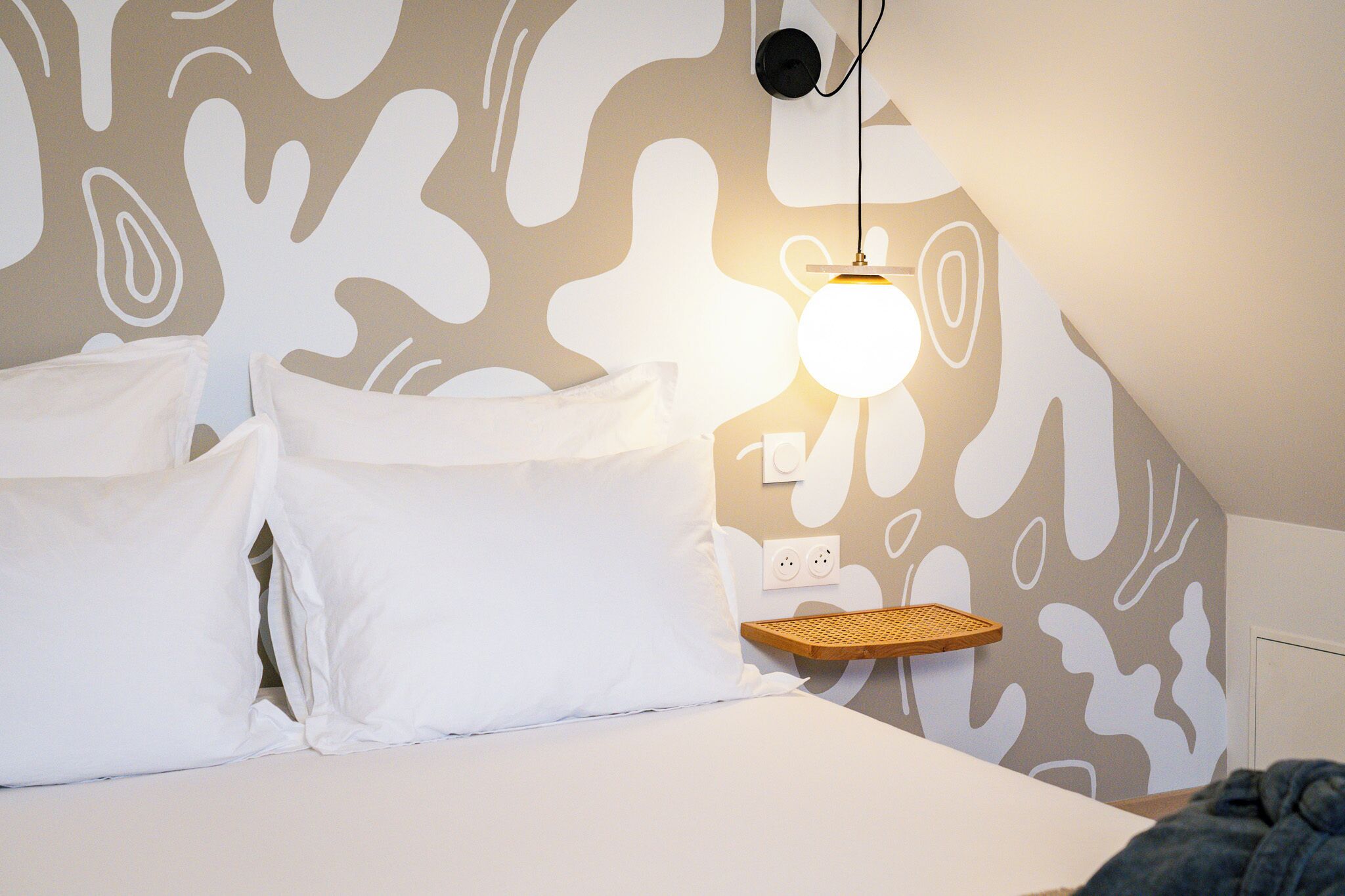 Un dormitorio con una cama, almohadas blancas, una lámpara de pared y un enchufe.