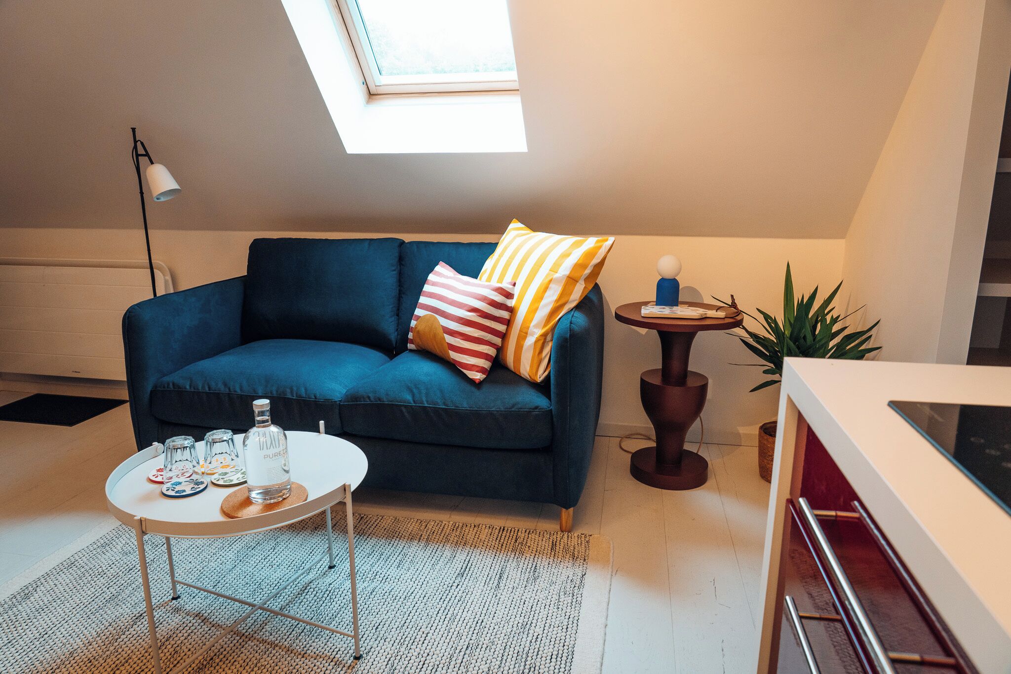 En moderne stue med en blå sofa, et rundt bord med drikkevarer, en gulvlampe og en potteplante.
