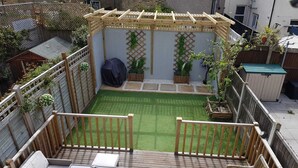 A small backyard with a wooden deck, a covered seating area, and a small garden with potted plants. - Cosy 4 bed house in great location. (London)