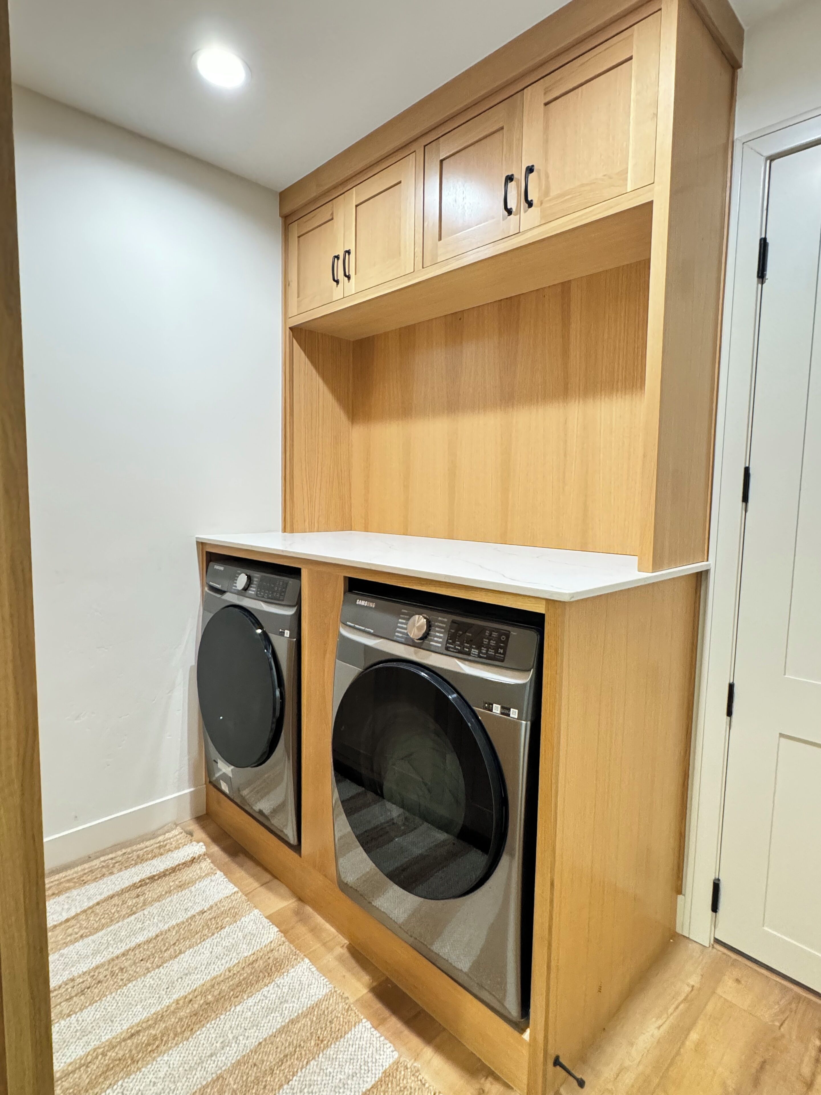 A laundry room with two front-loading washing machines, wooden cabinets above, and a white countertop.