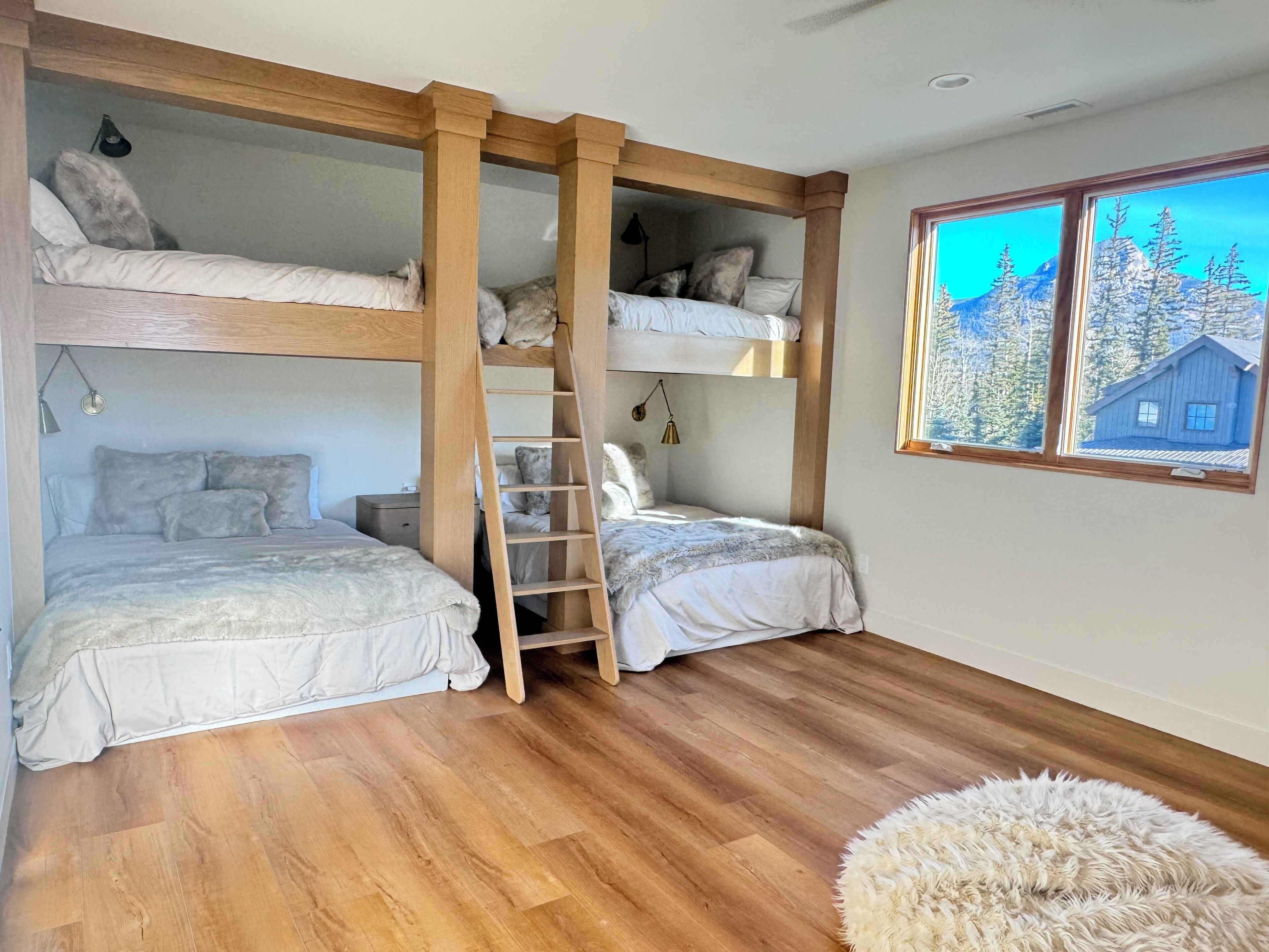 Quarto com beliche, escada de madeira, mesa de cabeceira e janela com vista para árvores e uma casa.
