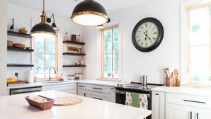 Une cuisine moderne avec des meubles blancs, un îlot central, des luminaires suspendus, une grande horloge murale et des étagères remplies d’ustensiles de cuisine.