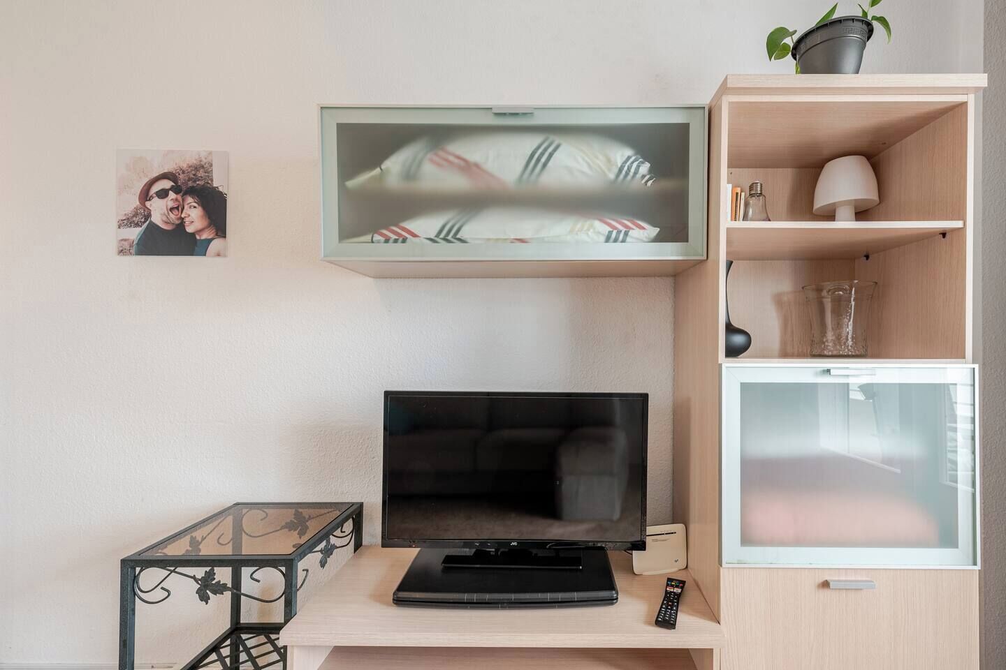 A living room with a flat-screen TV, a laptop, a remote control, a small table with a glass top, a potted plant, and a framed photo on the wall.