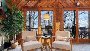 A cozy room with two upholstered chairs, a small round table, and a large window with a view of trees and a deck outside. - Cozy 6-bedroom cabin in Wintergreen Resort (Wintergreen Resort)
