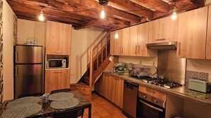 Eine rustikale Küche mit Holzausstattung, einem Edelstahlkühlschrank, einer Mikrowelle, einem Gasherd und einem Spülbecken. Ein Esstisch mit Stühlen und eine Treppe, die zu einer weiteren Ebene führen.