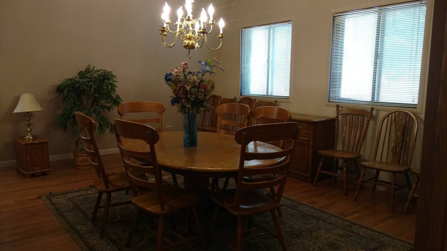 A dining room with a round wooden table, six wooden chairs, a chandelier, a vase with flowers, and two windows with blinds.