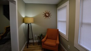 A corner room with a wooden floor, a corner chair, a floor lamp, a wall clock, and a window with blinds.