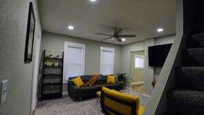 A living room with a sofa, a chair, a ceiling fan, and a TV mounted on the wall.