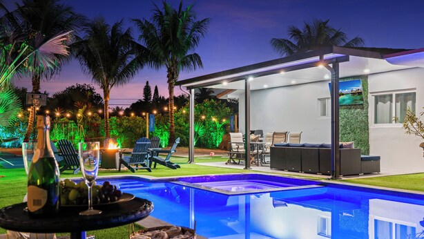 A modern outdoor pool area with a covered seating area, a table with bottles and glasses, and palm trees.