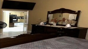 A bedroom with a bed, a dresser, a mirror, and a television mounted on the wall. - Luxe Escape Residential Housing (Portmore)