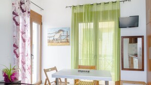A dining area with a table and chairs, a painting on the wall, a television mounted on the wall, and a window with curtains. - Single studio with Kitchen, 3 minutes (by foot) to the Beach & Hiking paths. (Diafani)