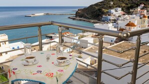 A balcony with a table set for two, overlooking a coastal town with buildings and a pier. - Single studio with Kitchen, 3 minutes (by foot) to the Beach & Hiking paths. (Diafani)