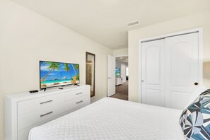 Quarto com cama, televisão sobre uma cômoda, porta e corredor que leva a outro espaço.