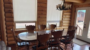 A wooden cabin interior with a round dining table, six chairs, a chandelier, and a large window.
