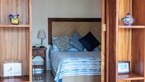 Un dormitorio con cama, almohadas, una mesita de noche, una lámpara y estantes de madera con objetos decorativos.