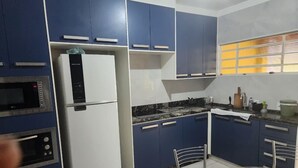 Een keuken met blauwe kastjes, een witte koelkast, een fornuis en een magnetron. Er staan twee stoelen en een tafel.