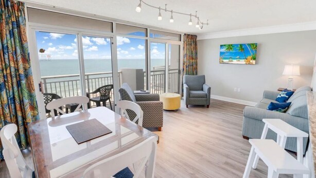 Une chambre avec un balcon donnant sur la mer, une table à manger, un canapé et un tableau au mur.