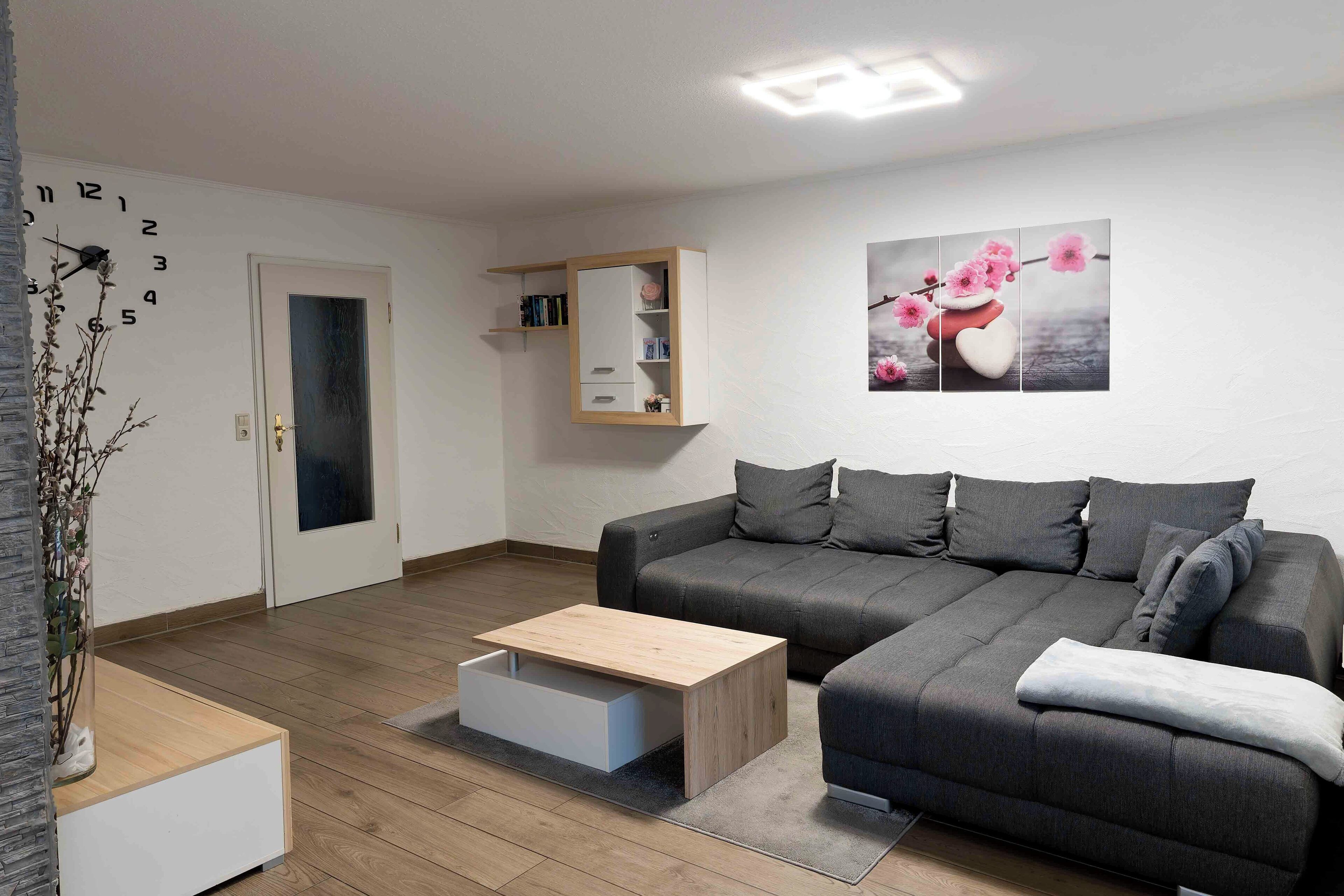 Ein modernes Wohnzimmer mit einem grauen Sofa, einem Couchtisch aus Holz mit Schublade, einem kleinen weißen Schrank und einer Wanduhr.
