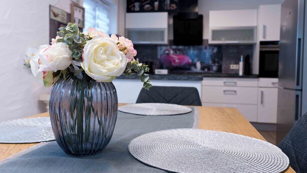 A modern kitchen with white cabinets, a black range hood, and a dining area with a wooden table and a vase of flowers. - Lake Constance pearl! Cozy 3.5 room apartment with beautiful garden near Lake Constance (Friedrichshafen)