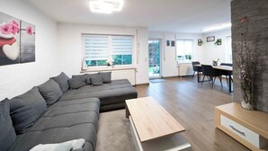 A modern living room with a grey sectional sofa, a wooden coffee table, and a dining area with a table and chairs. - Lake Constance pearl! Cozy 3.5 room apartment with beautiful garden near Lake Constance (Friedrichshafen)