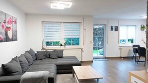 A modern living room with a grey sectional sofa, a wooden coffee table, and a dining area with a black chair and table. - Lake Constance pearl! Cozy 3.5 room apartment with beautiful garden near Lake Constance (Friedrichshafen)