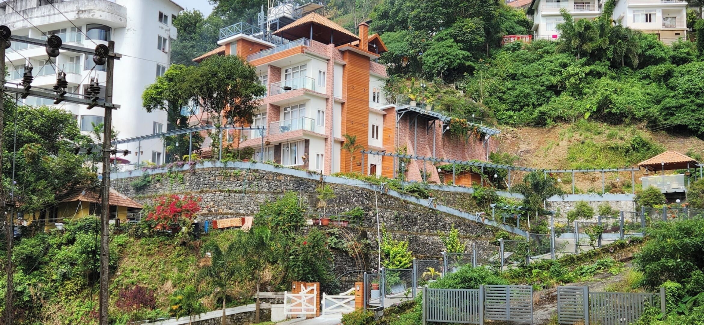 A hillside residential area with multi-story houses, a stone retaining wall, and a pedestrian bridge.