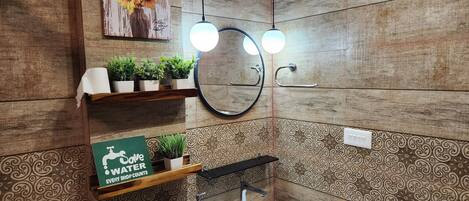 Un bagno con pareti rivestite in legno, un lavandino bianco, uno specchio, due mensole con piante e un cartello sulla conservazione dell'acqua.