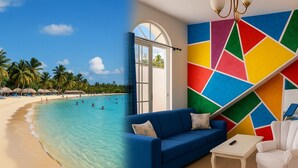 Ett rum med ett stort fönster med utsikt över en strand, en blå soffa, en vit fåtölj och en vägg med ett färgstarkt, geometriskt mönster.