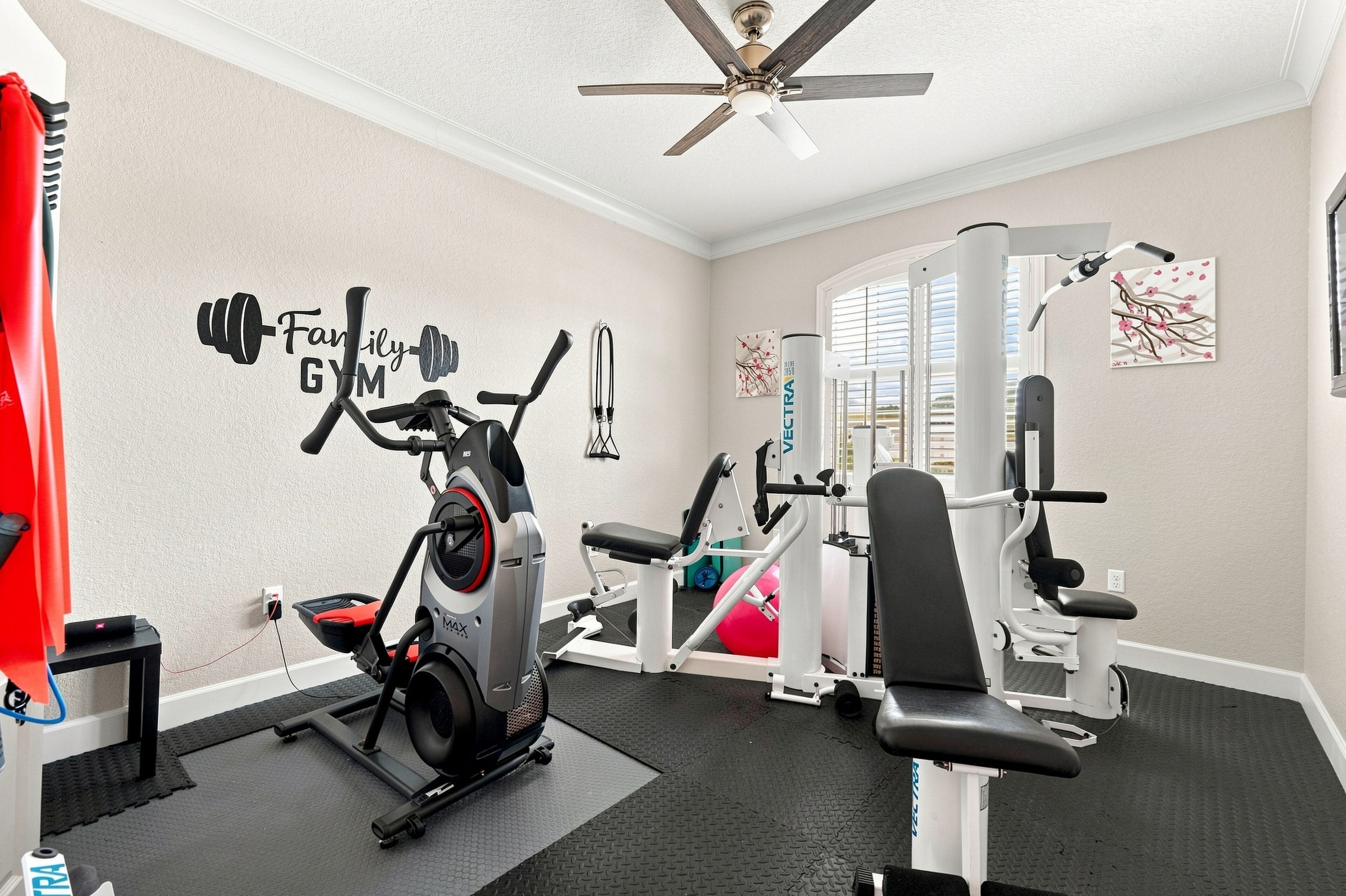 Un gimnasio en casa con equipo de ejercicio, un ventilador de techo y un adhesivo de pared motivacional.