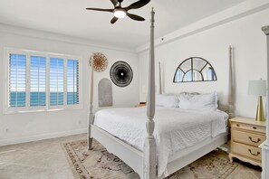 A bedroom with a four-poster bed, a ceiling fan, a large mirror, and a view of the ocean through the window.