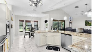 A modern kitchen with white cabinetry, granite countertops, and a dishwasher. There is a dining area with a table and chairs, and a large window providing a view of the outdoors. - Luxury Heated Pool • Game Room • Near Springs & Manatees (Beverly Hills)