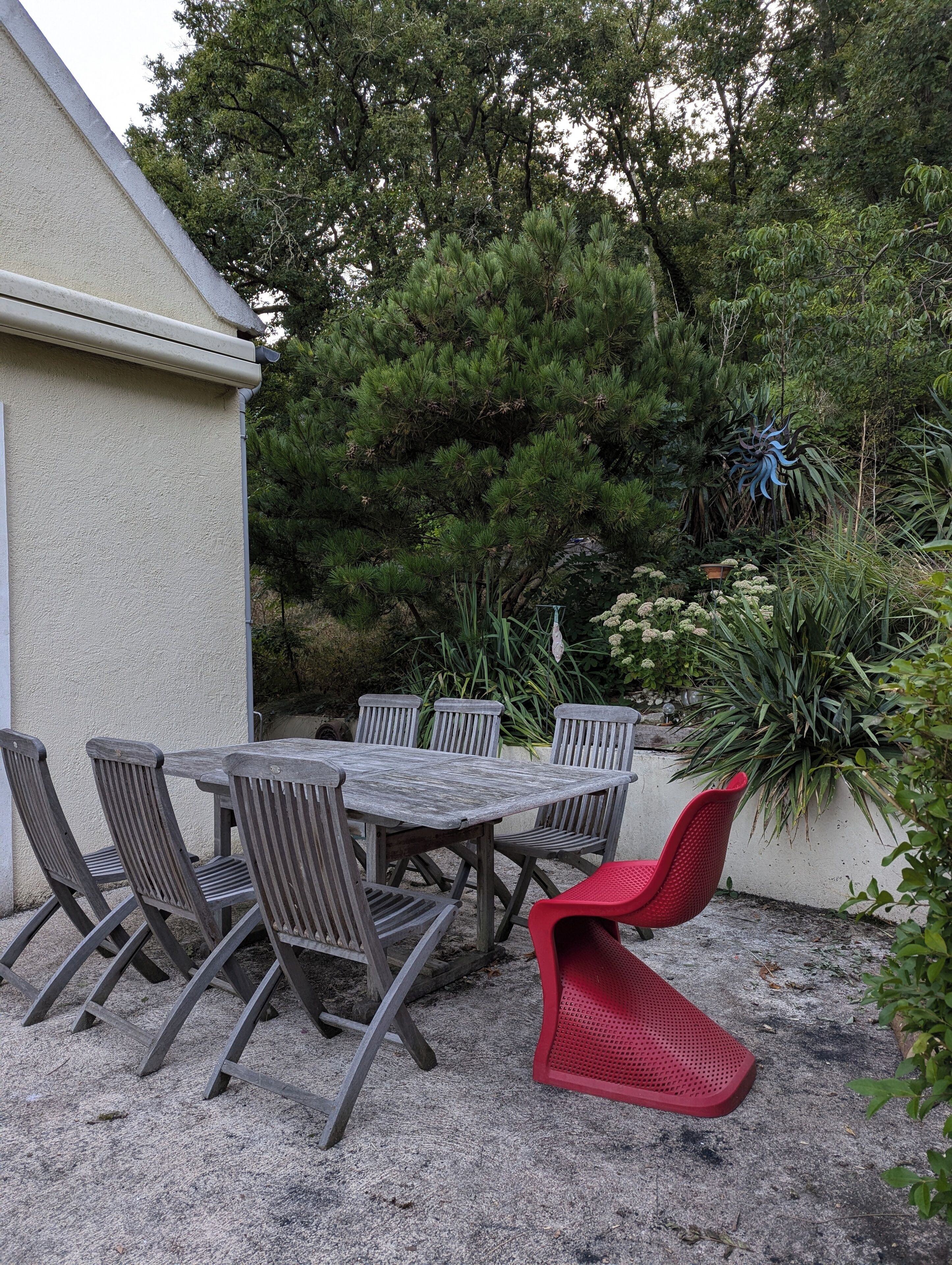 Eine Terrasse im Freien mit Tisch und Stühlen, umgeben von Grünpflanzen und einer Gebäudewand.