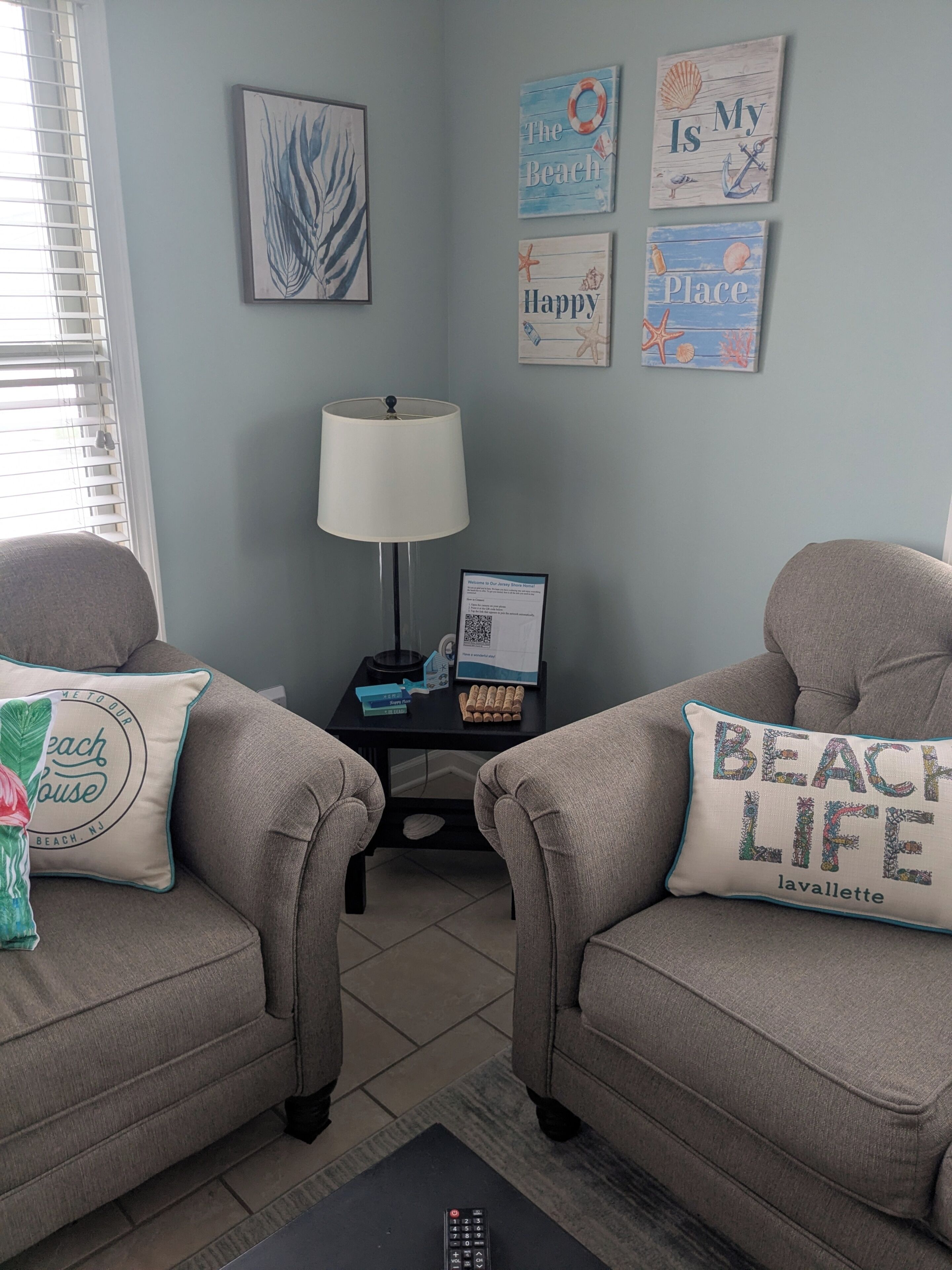 A living room with two armchairs, a side table with a lamp, a framed welcome sign, and beach-themed wall art.