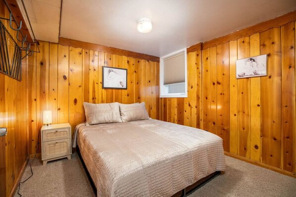 Un dormitorio con paneles de madera, una cama, una mesita de noche, una lámpara y dos cuadros colgados en la pared.