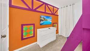 Une pièce avec un mur orange, une commode blanche, un tableau encadré et un téléviseur fixé au mur.