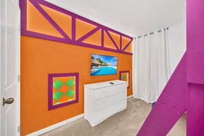 Une pièce avec un mur orange, une commode blanche, un tableau encadré et un téléviseur fixé au mur.