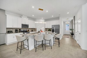 Een moderne keuken met witte kastjes, een centraal eiland met barkrukken, roestvrijstalen apparatuur en inbouwspots.