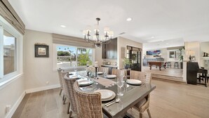 A spacious dining area with a long table set for six, surrounded by wicker chairs. A chandelier hangs above, and the room has a modern kitchen in the background.