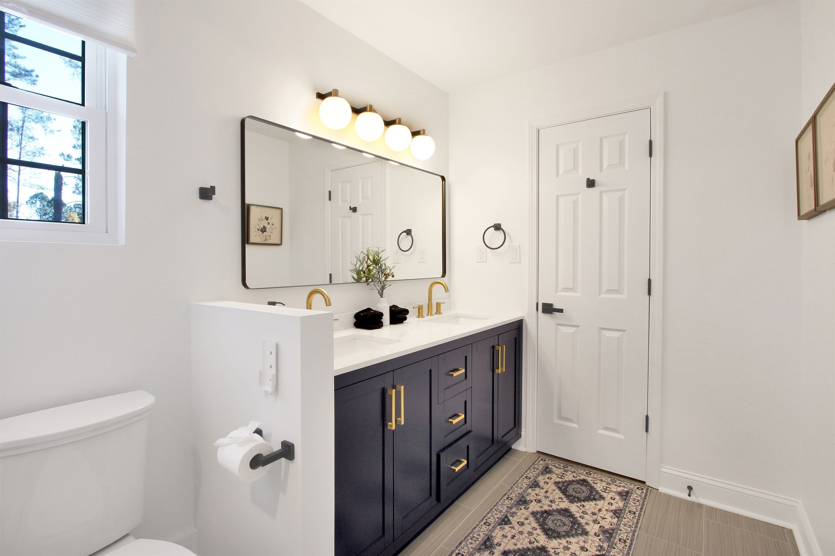 A bathroom with a white toilet, a black vanity with gold handles, a mirror with lights, and a patterned rug.