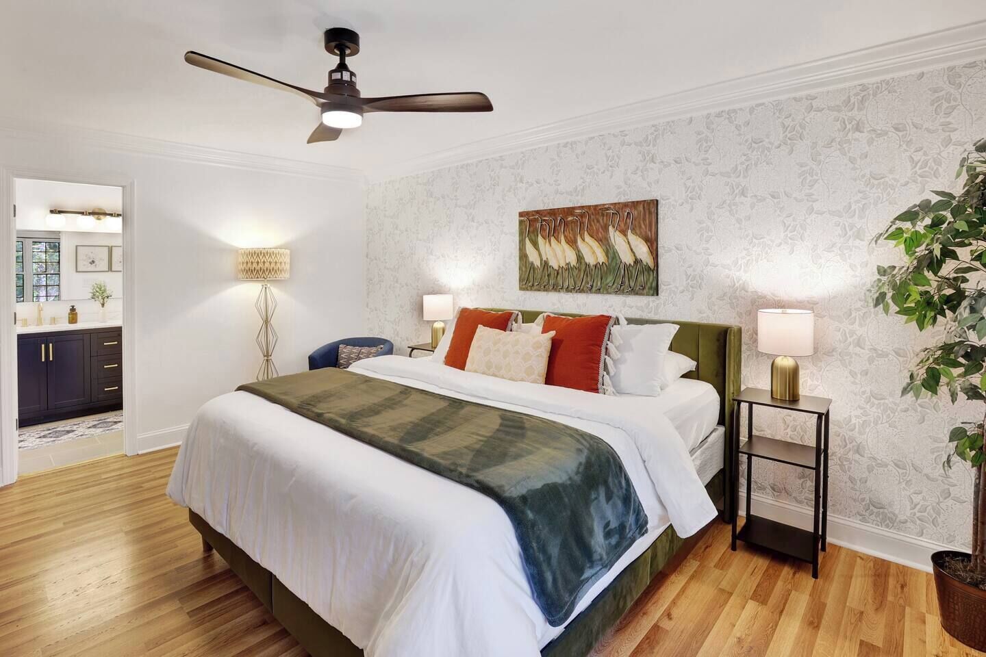 A bedroom with a bed, bedside lamps, a ceiling fan, a painting, and a potted plant.