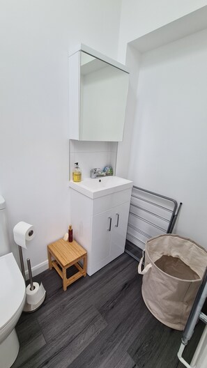 Une petite salle de bain avec un lavabo blanc, un petit tabouret en bois, un étendoir métallique et un panier à linge beige.