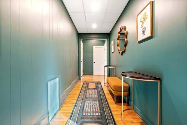 A hallway with teal walls, a wooden floor, a patterned rug, a bench, and a framed artwork on the wall.