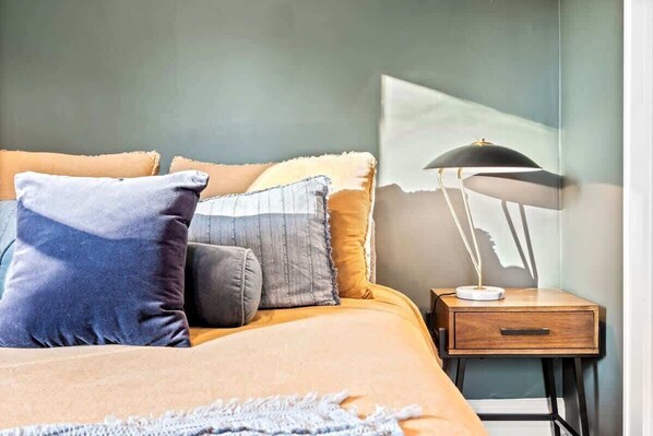 A neatly made bed with a variety of pillows, a bedside table with a lamp, and a wall with a partial view of a window.